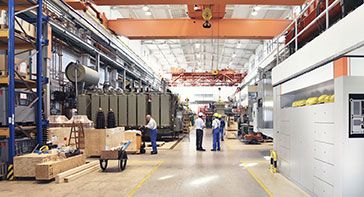 this picture is of a facility with overhead cranes and people with hardhats beneath them next to lumber and machinery and represents a manufacturing firm that does drug testing